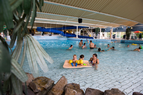 Overdekt zwembad bij een lodge met volwassenen en kinderen die spelen, en een blauwe glijbaan op de achtergrond.