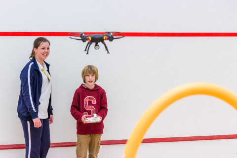 Jongen bestuurt een drone binnen in een lodge, terwijl een lachende vrouw toekijkt naast hem.
