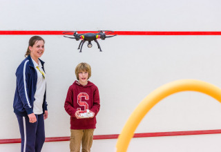 Jongen bestuurt een drone binnen in een lodge, terwijl een lachende vrouw toekijkt naast hem.