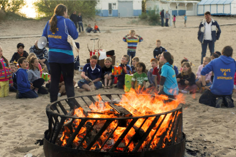 Kinderen en volwassenen verzamelen zich rond een groot kampvuur op het strand bij een lodge, met begeleiding.