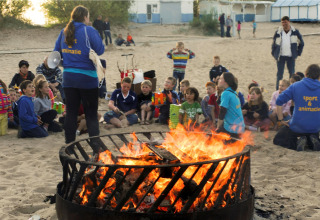 Kinderen en volwassenen verzamelen zich rond een groot kampvuur op het strand bij een lodge, met begeleiding.