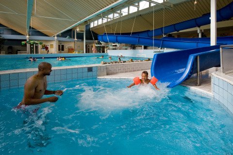 Overdekt zwembad bij een lodge met kinderen op een blauwe glijbaan en volwassenen die toezicht houden.