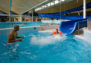 Overdekt zwembad bij een lodge met kinderen op een blauwe glijbaan en volwassenen die toezicht houden.