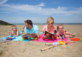 Een gezin ontspant op het strand met tijdschriften, fruit en ijsjes, omringd door speelgoed en strandspullen.