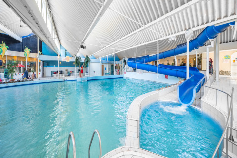 Binnenzwembad in een lodge met blauwe glijbaan, hoog wit plafond en tropische decoratie.