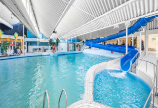 Binnenzwembad in een lodge met blauwe glijbaan, hoog wit plafond en tropische decoratie.