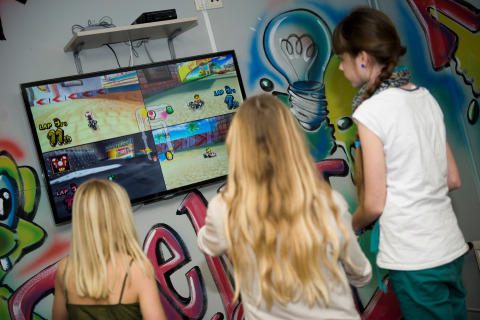 Drie kinderen spelen samen een videogame op een groot tv-scherm in een kleurrijke, met graffiti versierde lodge.