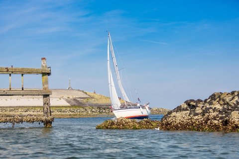 Zeilboot vaart dicht bij rotsen en een oude houten steiger aan een zandige kust onder een blauwe lucht.
