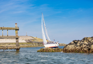 Zeilboot vaart dicht bij rotsen en een oude houten steiger aan een zandige kust onder een blauwe lucht.