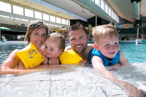 Blije familie samen in het binnenzwembad van een lodge, kinderen dragen zwembandjes en zwemvest.