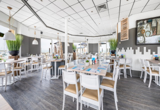 Lichte, moderne lodge-restaurant met witte stoelen, houten vloer, grote ramen en groene kamerplanten.