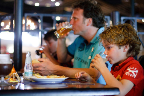 Een gezin eet en drinkt samen in een lodge, met een man die bier drinkt en een kind aan tafel.