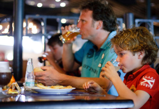 Een gezin eet en drinkt samen in een lodge, met een man die bier drinkt en een kind aan tafel.
