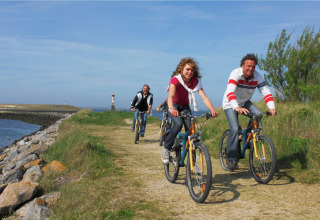 Drie personen fietsen over een pad langs het water met groen gras en een blauwe lucht op een zonnige dag.