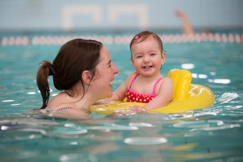 Een vrouw en een lachende baby in een geel zwemzitje genieten samen in het binnenzwembad van een lodge.
