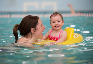 Een vrouw en een lachende baby in een geel zwemzitje genieten samen in het binnenzwembad van een lodge.
