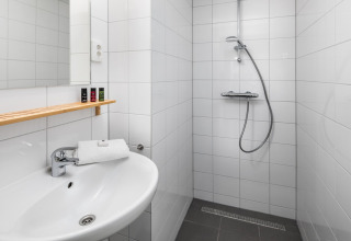 Moderne lille badeværelse i et tiny house med hvidt fliseindretning, bruser, håndvask og træhylde.