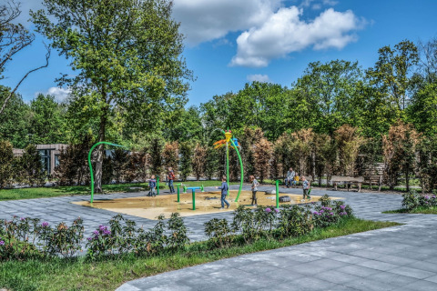 Kinder spielen auf einem Wasserspielplatz neben einem Tiny House, umgeben von Bäumen und Himmel.