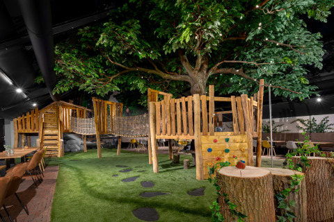Aire de jeux intérieure avec cabane dans les arbres, pont en corde, pelouse artificielle et grandes tables.