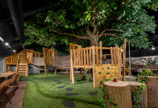 Indoor playground with wooden treehouse, rope bridge and fake grass, next to tables and chairs under a big tree.