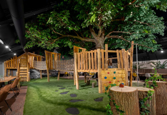 Aire de jeux intérieure avec cabane dans les arbres, pont en corde, pelouse artificielle et grandes tables.