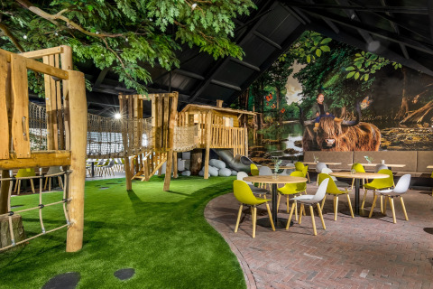 Espace de jeux intérieur avec cabanes en bois, gazon artificiel et chaises modernes jaunes et grises.