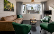Salon lumineux dans une tiny house avec canapé marron, fauteuils verts, tables rondes et grandes fenêtres.