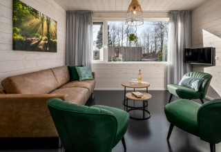 Bright living area in a tiny house with a brown sofa, green chairs, round tables, and large windows.