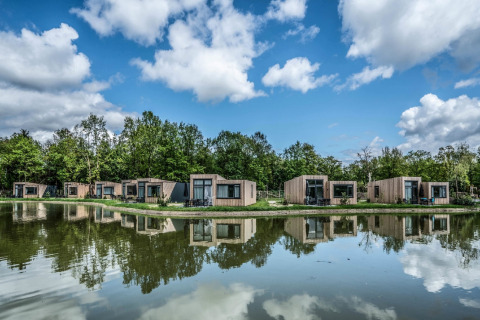 Modern tiny houses lined up by a tranquil pond, reflecting the sky and trees, with lush greenery behind.