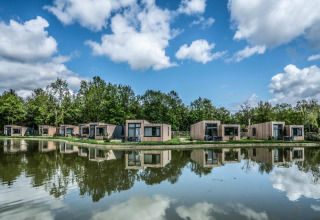 Modern tiny houses lined up by a tranquil pond, reflecting the sky and trees, with lush greenery behind.