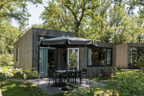 Modern tiny house Holiday Park «Schaijk 2» in the Netherlands with patio, chairs, umbrella, and greenery.