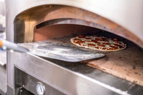 A pizza is being placed into a stone oven using a pizza peel in a tiny house kitchen setting.