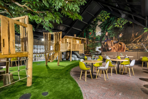 Indendørs legeplads og café-område med træhus, klatrebro, kunstgræs og farverige stole og borde.
