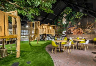 Indoor play area with wooden treehouse structure, rope bridge, artificial grass, tables and colorful chairs.