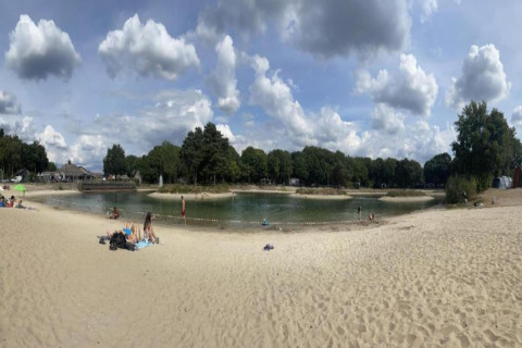 Panoramic view of a small lake with sandy beach and trees, as seen from a tiny house under blue sky.