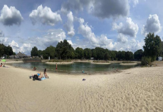 Vista panoramica di un piccolo lago con spiaggia sabbiosa e alberi, scattata da una tiny house sotto un cielo azzurro.