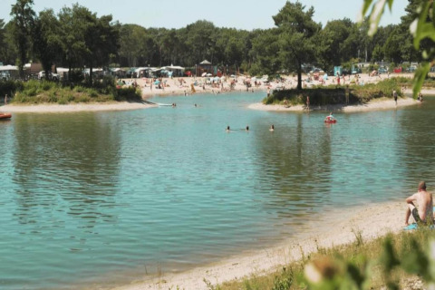 Vue sur un lac où des gens nagent, plage de sable, arbres et petites maisons à l’arrière-plan.