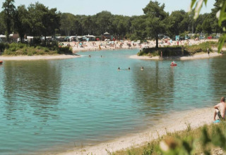 Vue sur un lac où des gens nagent, plage de sable, arbres et petites maisons à l’arrière-plan.