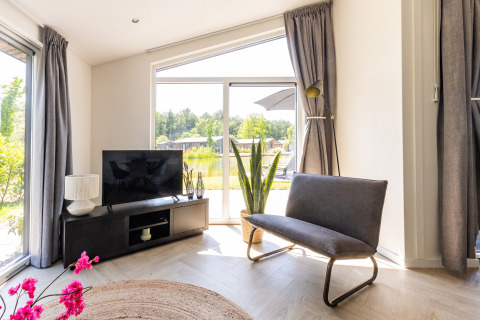 Salon moderne avec télévision, banc gris, plante et grandes baies vitrées au chalet Holiday Park Molenvelden 8.