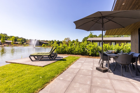 Patio at Holiday Park Molenvelden 8 in the Netherlands, with loungers, umbrella, lake, and fountain view.