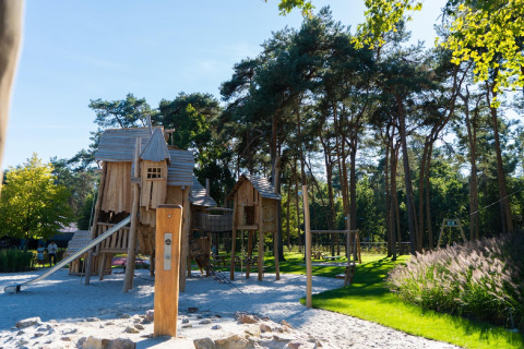Zona de juegos de madera cerca de un chalet, rodeada de árboles y césped en un entorno natural.