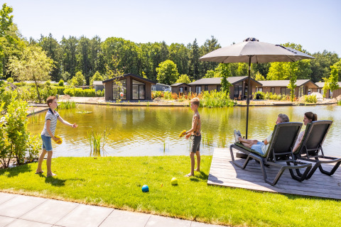 Family relaxes by a pond with chalets at Holiday Park Molenvelden 8, Netherlands, enjoying a sunny day.