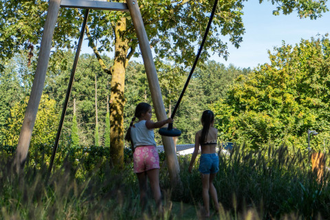 Due ragazze giocano con un’altalena tra l’erba alta vicino a un chalet immerso tra gli alberi verdi.