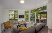 Lichte lodge woonkamer met moderne meubels, grote ramen en uitzicht op terras en omliggend bos.