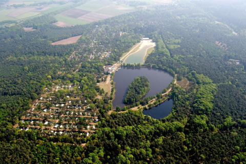 Aerial view of a lodge by a lake, surrounded by dense forest, cabins, and open green fields.