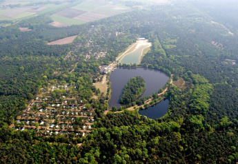 Aerial view of a lodge by a lake, surrounded by dense forest, cabins, and open green fields.