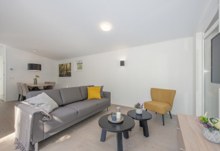 Stilfuldt opholdsområde i en lodge med grå sofa, gul lænestol, runde borde og spiseplads bagved.
