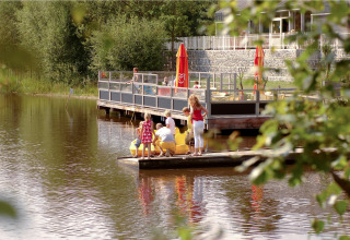 Familie nyder tid ved vandet nær glampingindkvartering med terrasse og røde parasoller i baggrunden.