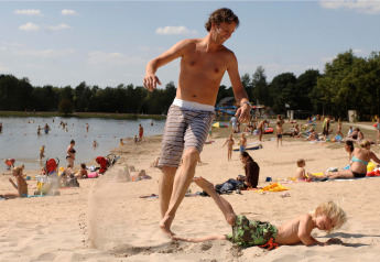 Vater und Kind spielen am Sandstrand eines Campingplatzes mit vielen Menschen und sonnigem Wetter.