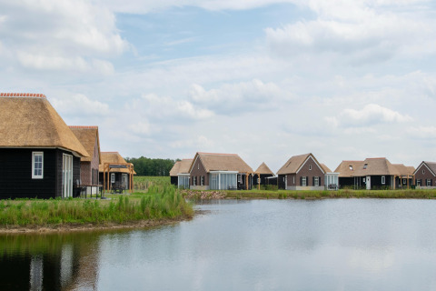 Glamping-Hütten mit Strohdächern am See in ländlicher Umgebung unter bewölktem Himmel.